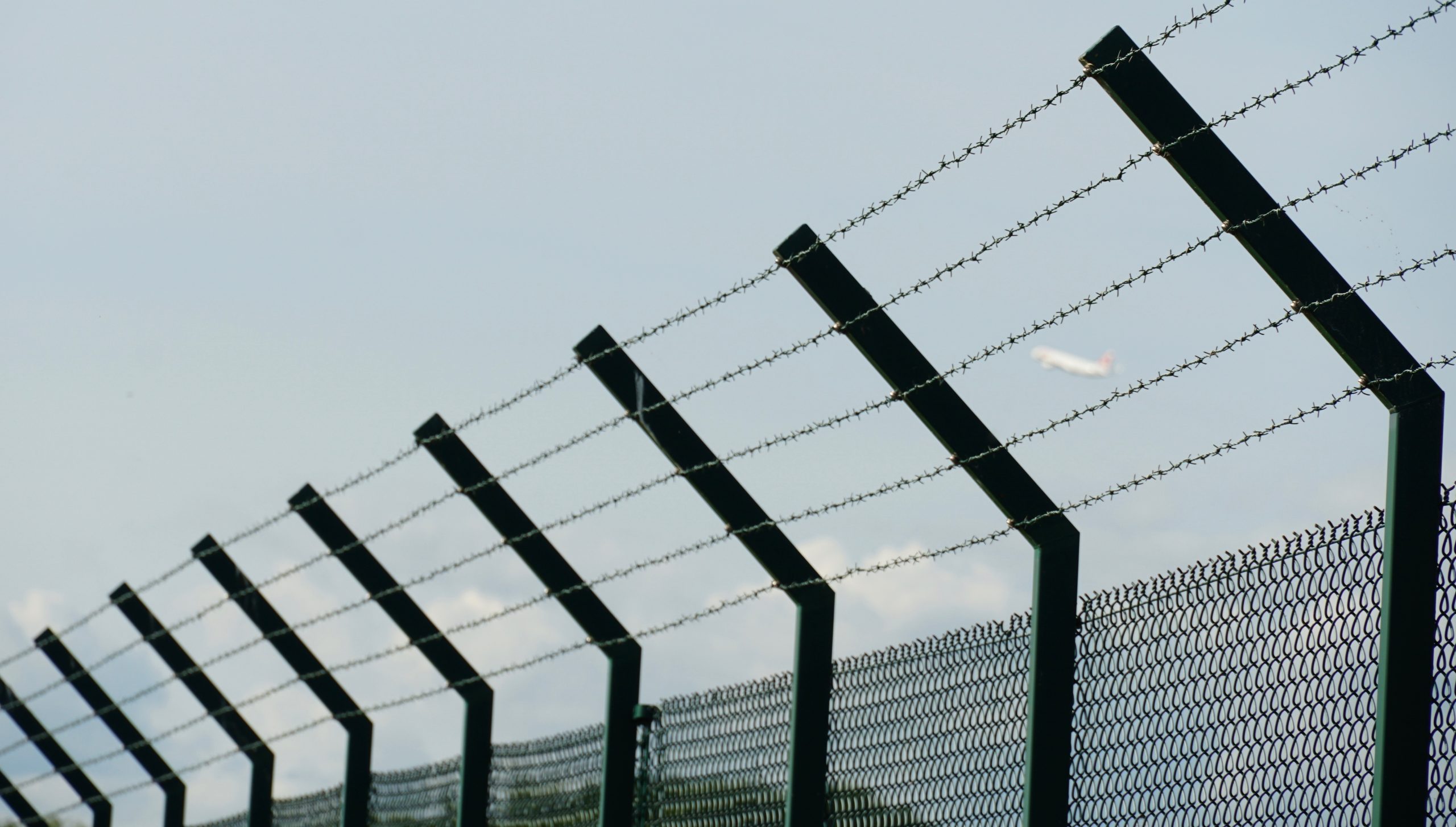 A tall fence around the ground of Zurich Airport made of woven wire and enhanced with barbed wire as protection from unauthorized entry. There is a vague silhouette of a plane on the background.